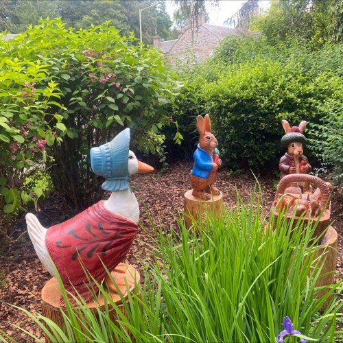 In the Beatrix Potter Garden Trail Dunkeld, Jemima PuddleDuck and Peter Rabbit gather around a picnic basket. Carved from wood and brightly painted these creatures nestle into a small space between hedgerow and pathway